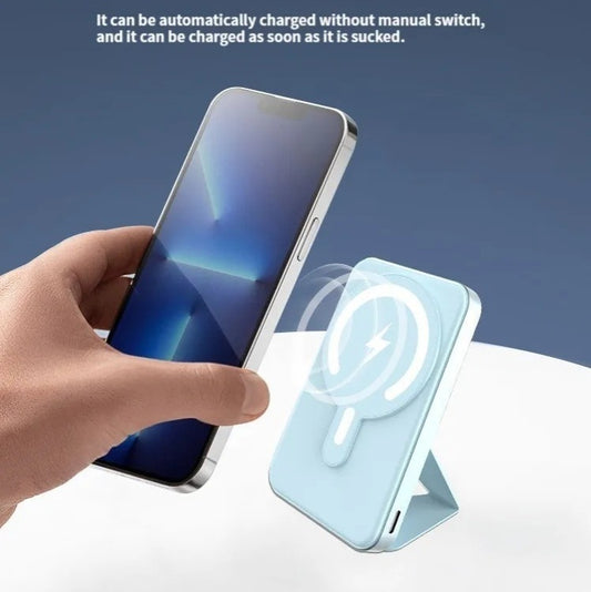 Mano sujetando iPhone acercándolo a powerbank magnético azul claro con soporte plegable y símbolo de carga MagSafe